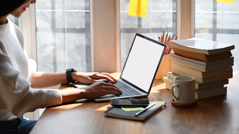 A person types on a laptop, perhaps planning to start a blog, at a wooden desk. Nearby are stacked books, a smartphone, a notebook, a cup of coffee, and pencils in a holder. Sunlight streams through large windows, casting a warm glow on the workspace.